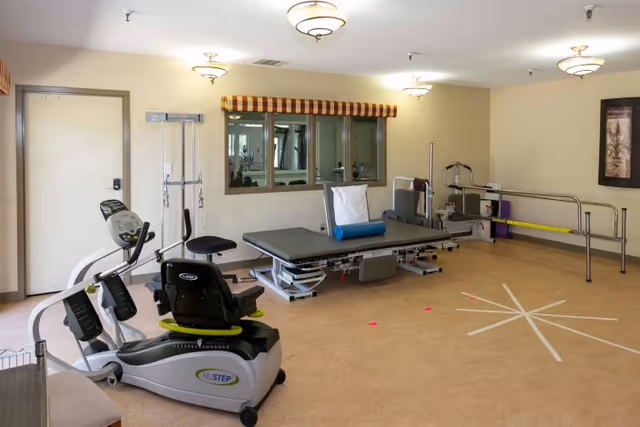 A rehabilitation room with exercise equipment including a NuStep recumbent cross trainer, a padded therapy table with a blue bolster, parallel bars for walking exercises, and a chair. The room has beige walls, ceiling lights, a window with a striped valance, and a framed picture on the wall.
