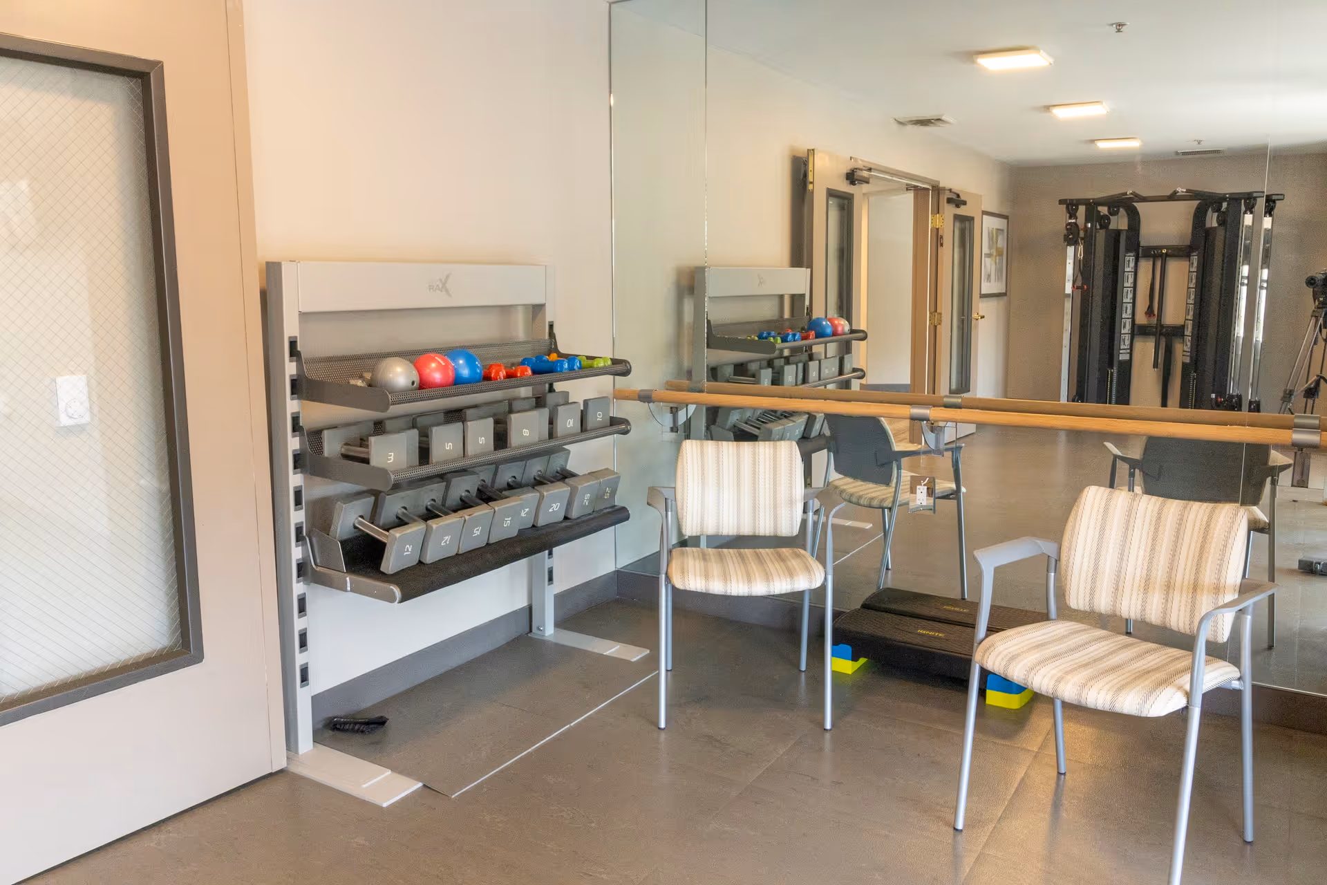 A fitness room with a rack of various dumbbells and exercise balls, two striped cushioned chairs, a ballet barre mounted on a mirrored wall, and exercise equipment in the background.