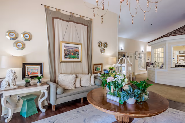 A cozy living room area with a beige sofa adorned with cushions, a wooden round table in the center decorated with various green plants and flowers, a side table with a white lion statue, lamp, and framed pictures on the wall. The room has soft lighting and a welcoming atmosphere with a reception area visible in the background.