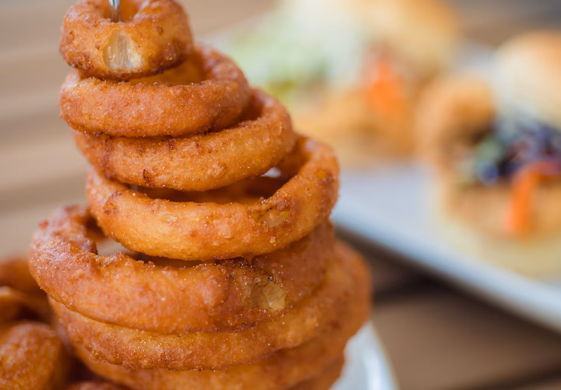 Close-up view of a stack of crispy golden-brown onion rings on a plate with blurred sandwiches in the background.