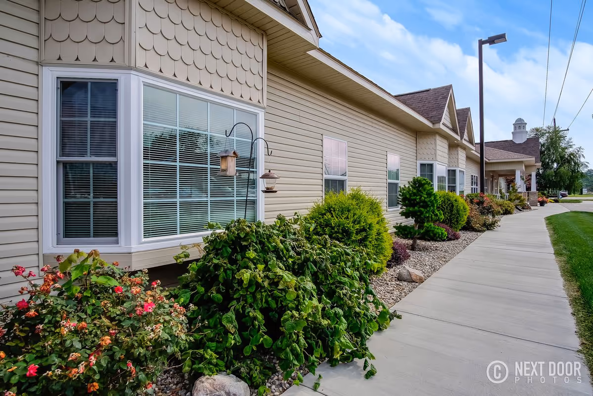 Exterior view of a single-story assisted living building with windows, landscaped shrubs, and a sidewalk.