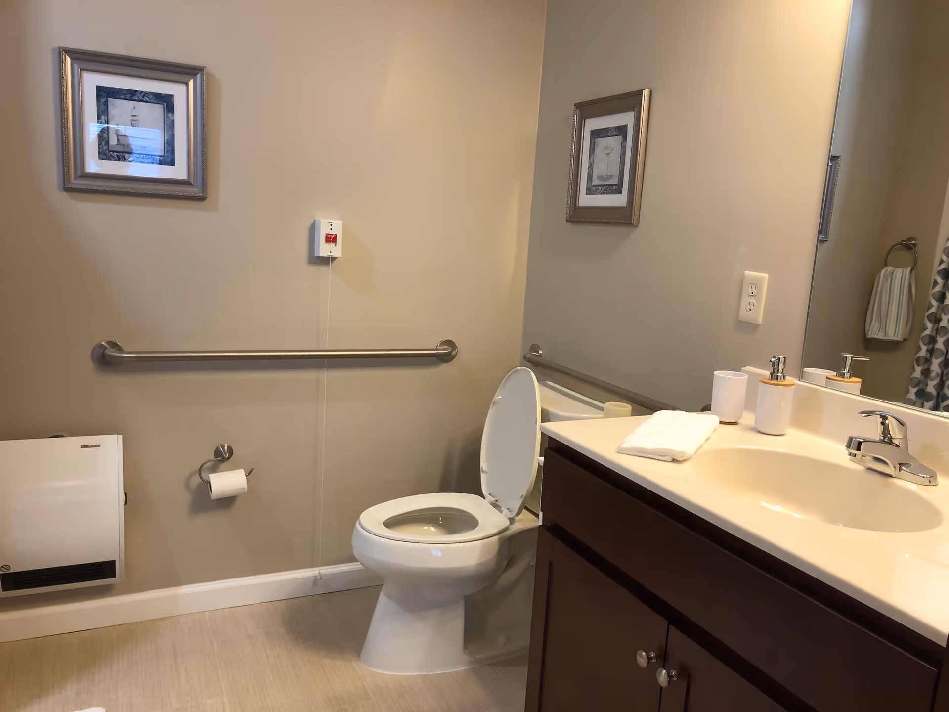 A clean bathroom with a white toilet with the lid open, a beige countertop with a sink, soap dispenser, and a cup. There are two framed pictures on the beige walls, a metal grab bar along the wall behind the toilet, a toilet paper holder with a roll of toilet paper, a wall-mounted heater, and a towel hanging on a ring near a shower curtain.