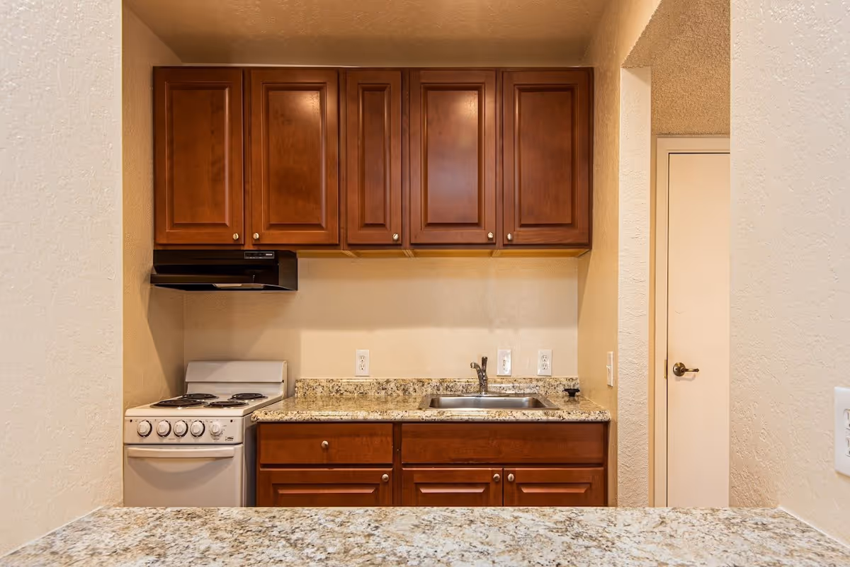 Small kitchen area with wooden cabinets, a granite countertop, a white electric stove with four burners, a stainless steel sink, and a small door to the right.