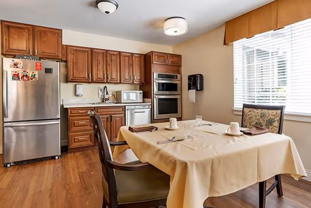 A kitchen and dining area with wooden cabinets, stainless steel refrigerator, built-in oven, microwave, dishwasher, and a dining table covered with a beige tablecloth set with plates, cups, and utensils. There is a window with blinds and a valance letting in natural light.