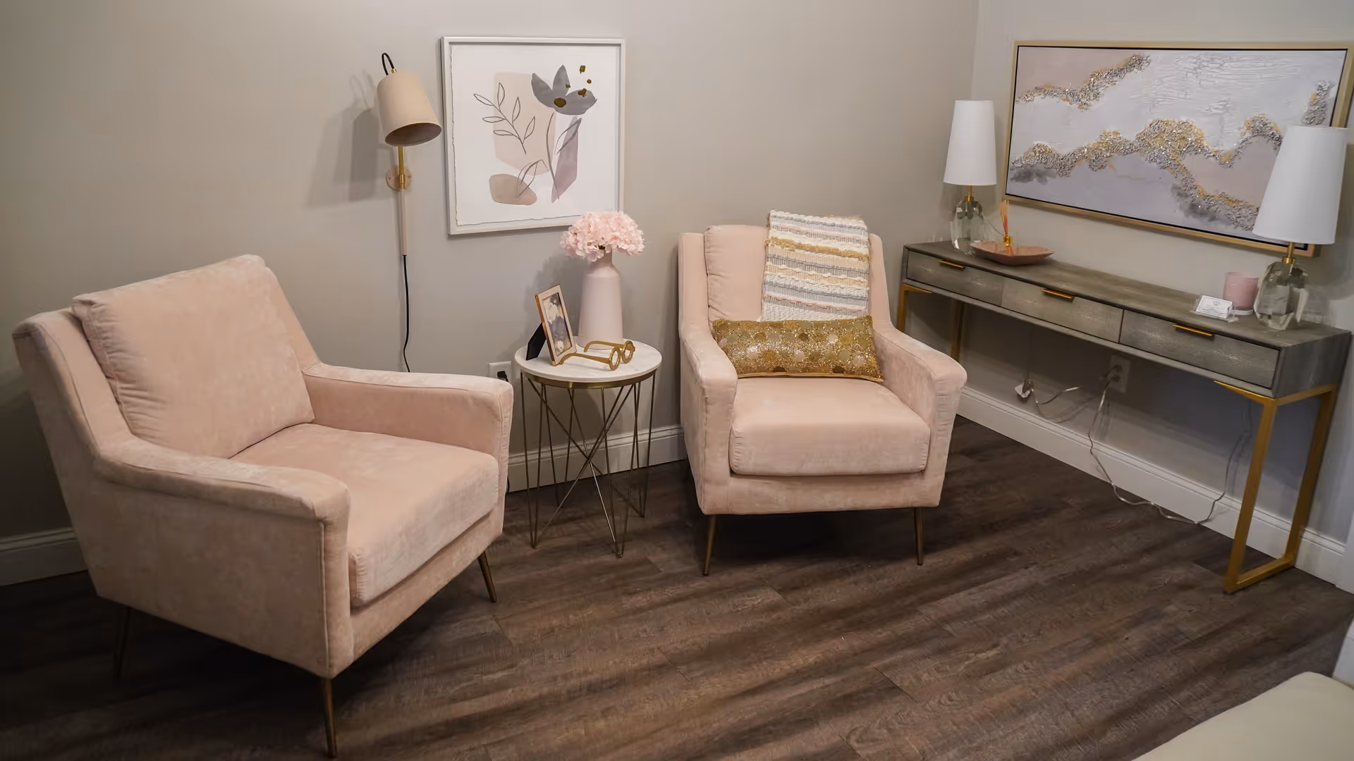 A cozy seating area with two blush pink armchairs separated by a small round side table holding a vase with pink flowers and a framed photo. A wall-mounted lamp is above the table. To the right, there is a long console table with two lamps, decorative items, and a large abstract painting hanging above it. The room has wooden flooring and light-colored walls.