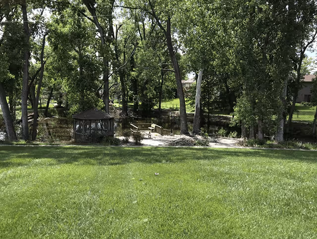 A grassy outdoor area with tall trees surrounding a small wooden gazebo and a wooden dock extending over a pond or small body of water. The scene is peaceful and shaded by the trees.