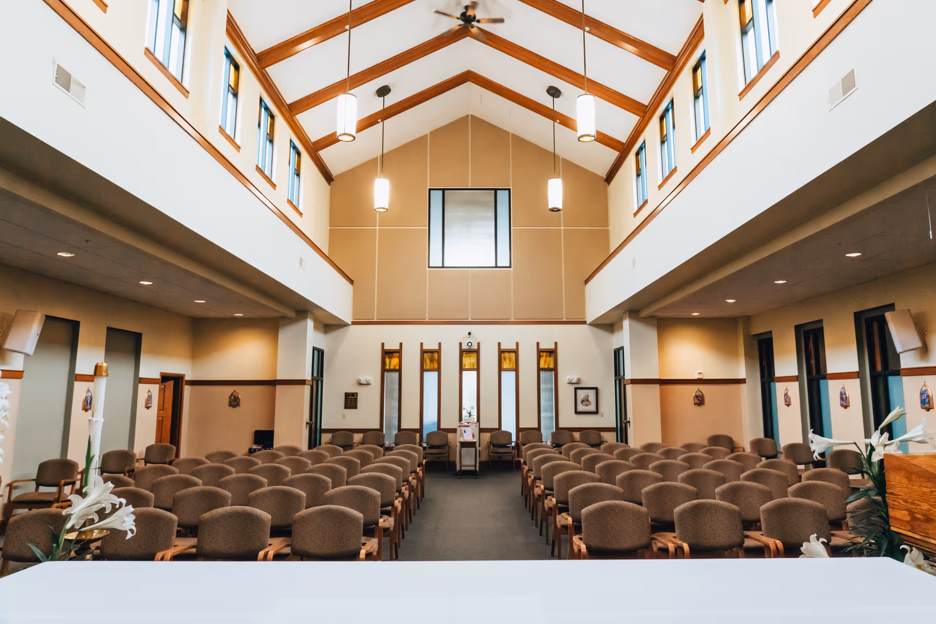 Interior view of a chapel or large meeting room with rows of brown cushioned chairs arranged facing a podium at the front. The room has high vaulted ceilings with wooden beams, hanging pendant lights, and tall windows near the ceiling letting in natural light. There are decorative flowers near the podium and framed pictures on the walls.