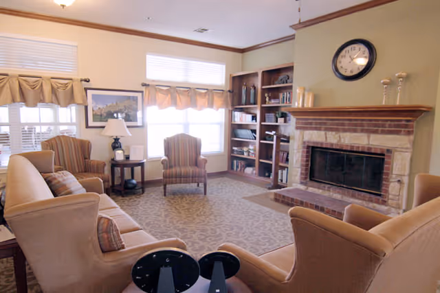 A cozy community living room with sofas and armchairs arranged around a brick fireplace and built-in bookshelves beneath windows.