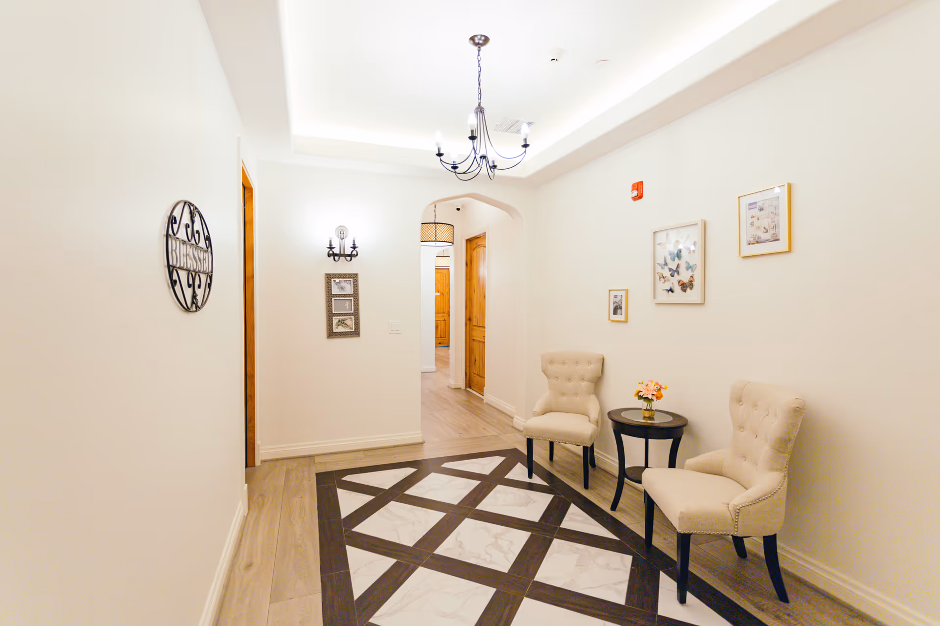 Bright interior hallway seating area with two upholstered chairs, a small round table, framed artwork, and a chandelier.
