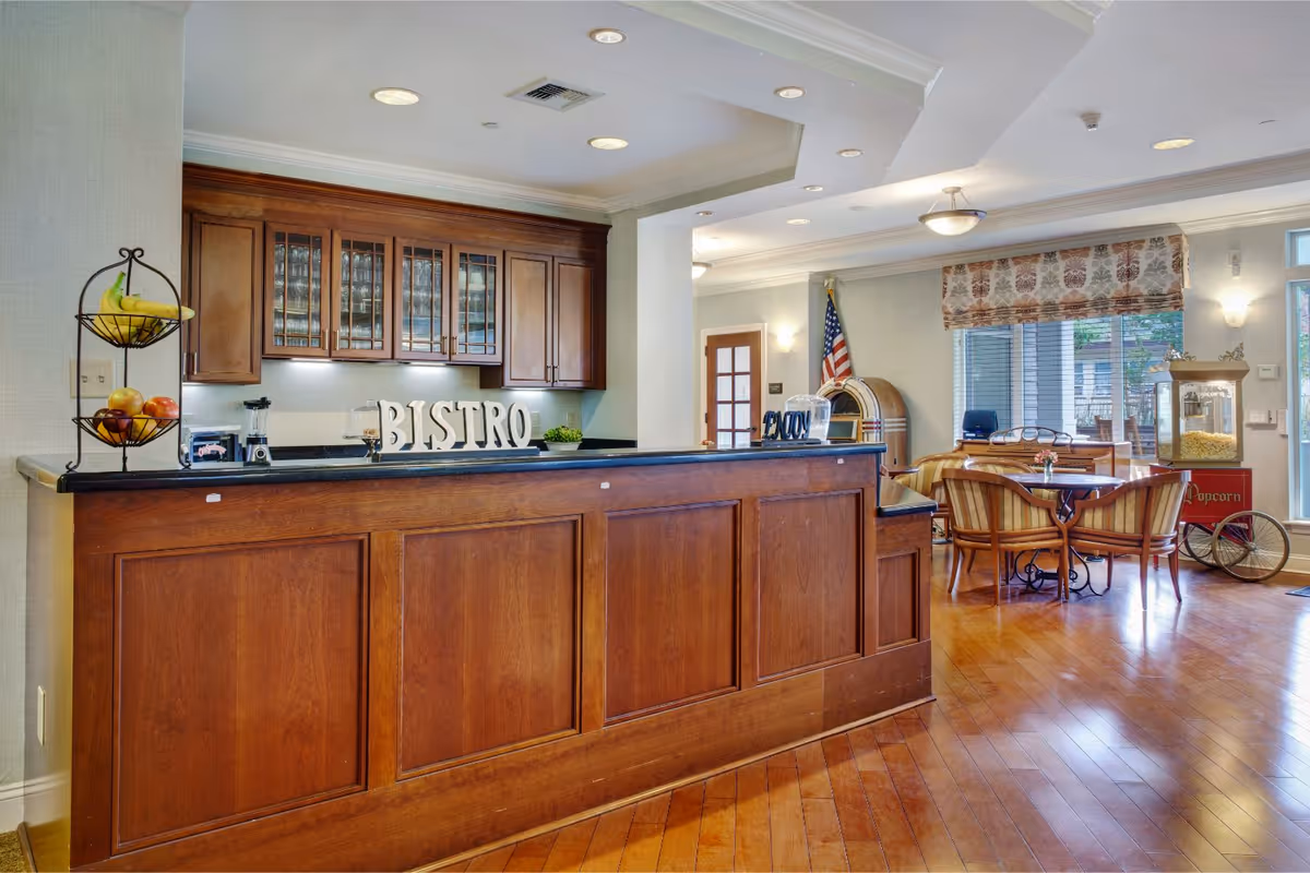 Interior view of a senior living facility's bistro area featuring a wooden counter with the word 'BISTRO' displayed on it, wooden cabinets with glass doors behind the counter, a fruit stand with bananas and apples, a seating area with round tables and chairs, an American flag, a popcorn machine, and large windows with patterned curtains allowing natural light to fill the room.