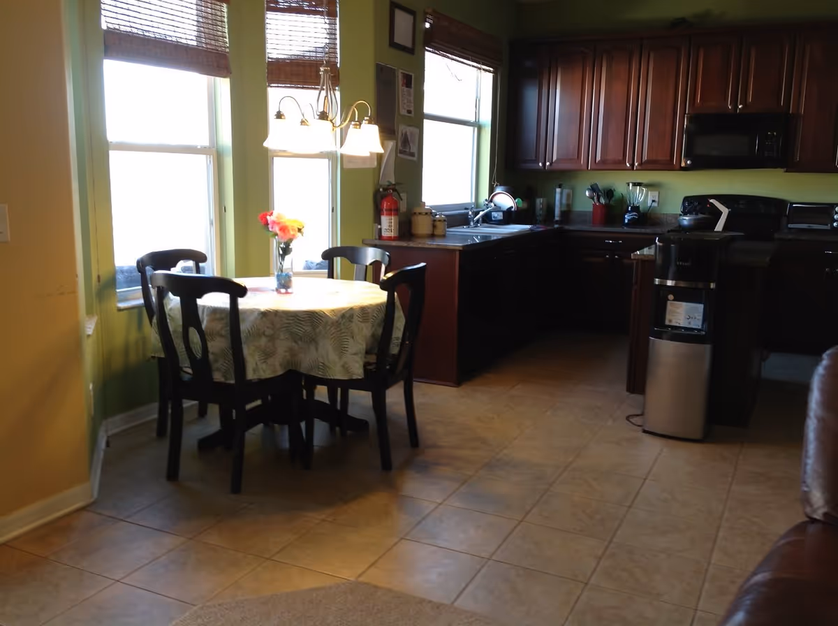 A kitchen and dining area with a round table covered with a patterned tablecloth and four dark wooden chairs. A vase with colorful flowers is placed on the table. The kitchen has dark wooden cabinets, a sink under a window, and various appliances including a microwave, toaster oven, and a water dispenser. The walls are painted green and there are three windows with brown blinds. The floor is tiled.