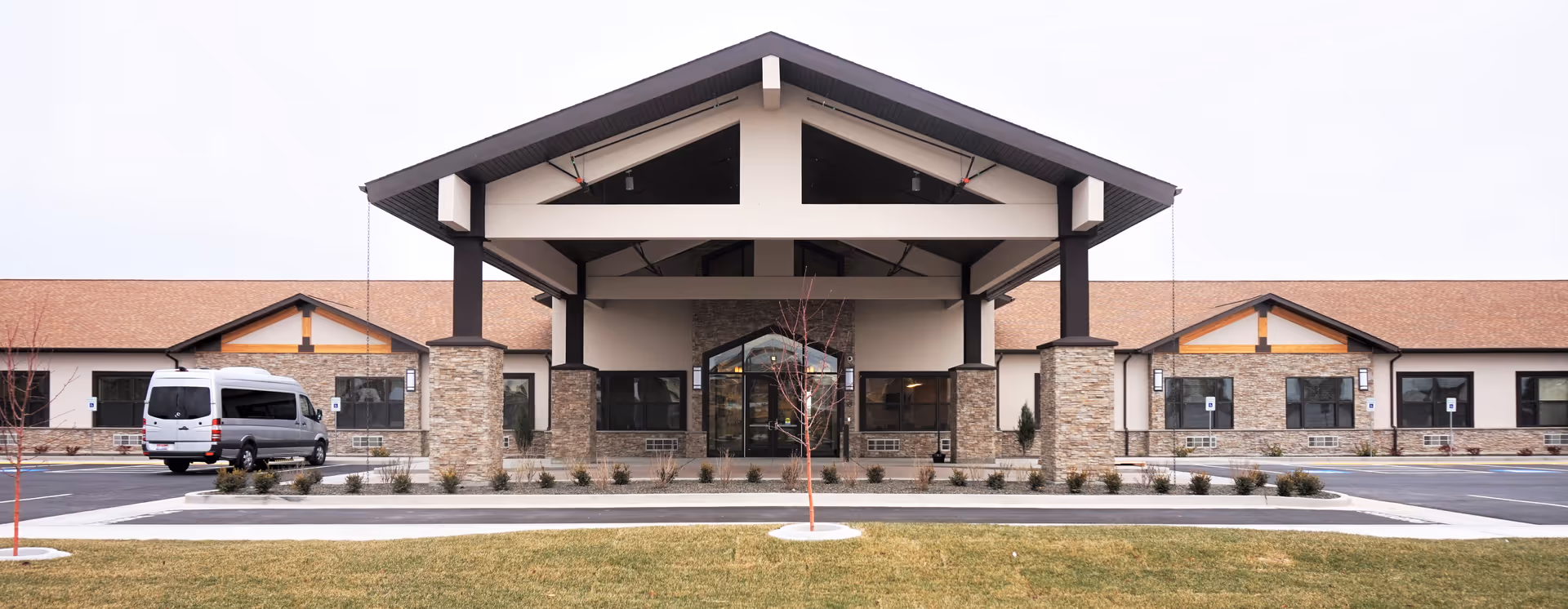 Front exterior of a single-story senior living facility with a covered entrance supported by stone pillars and a parked van.