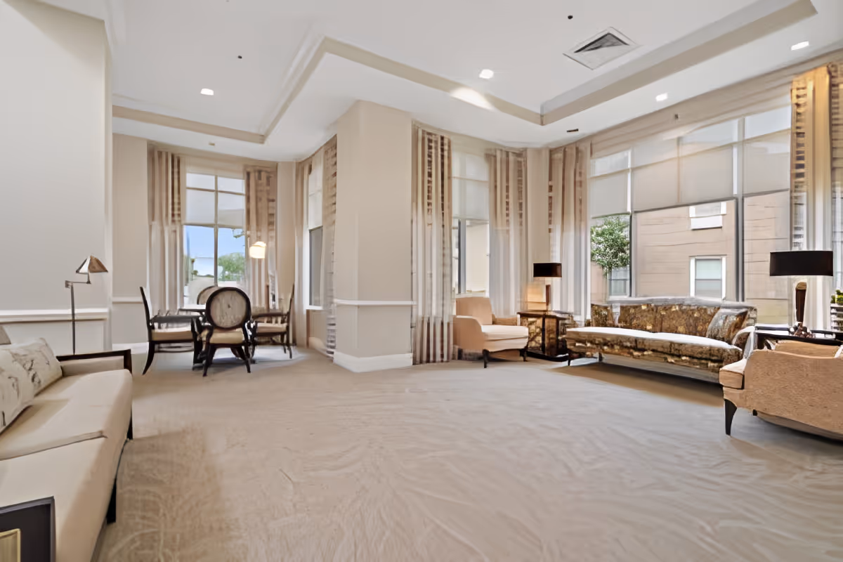 A spacious, well-lit living room area with beige carpeting and cream-colored walls. The room features multiple seating options including a beige sofa, two armchairs, and a patterned couch. There is a small round table with four chairs near large windows covered with sheer curtains, allowing natural light to fill the space. Two table lamps are placed on side tables beside the seating area.