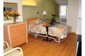 A bright bedroom in a care facility with an adjustable hospital-style bed, wooden dresser, chair, bedside table, and floral arrangements.