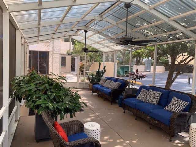 A covered outdoor patio area with a transparent roof, featuring wicker furniture with blue cushions and patterned pillows. There are ceiling fans mounted on the roof, several potted plants, and a small white stool. The patio overlooks a courtyard with trees and a building in the background.