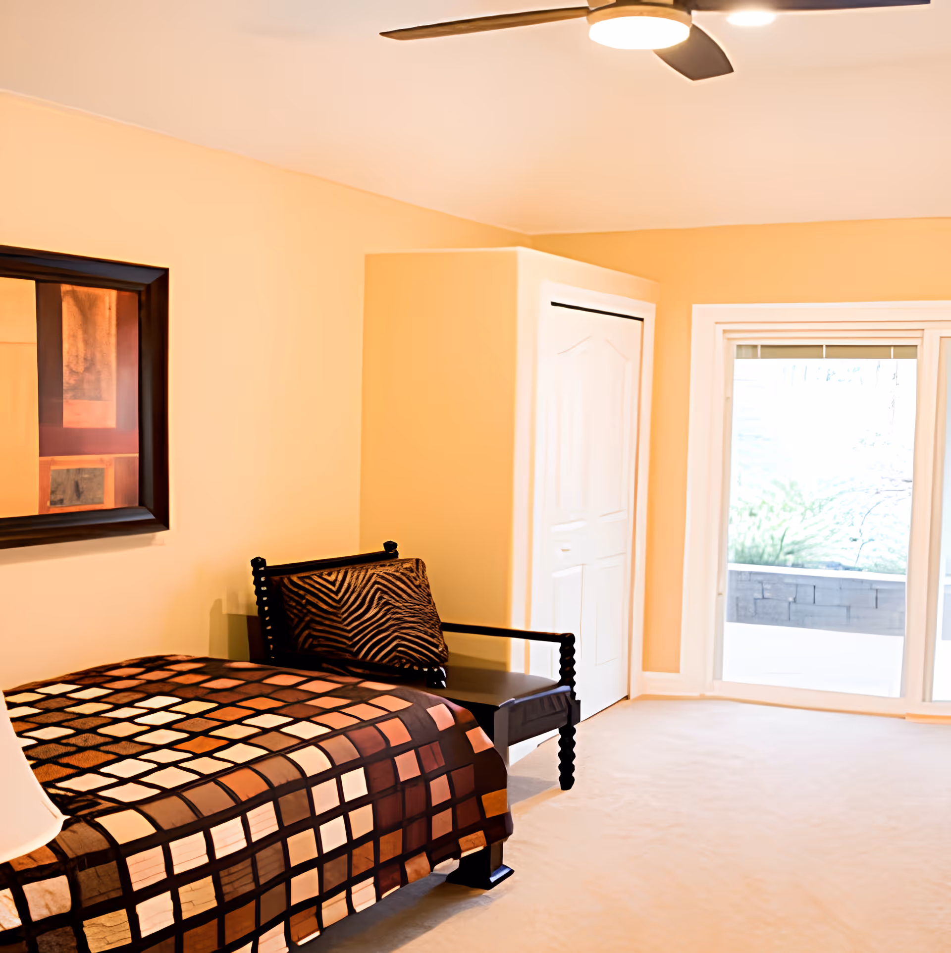 A bedroom with a bed covered in a brown and beige checkered blanket, a black wooden chair with a patterned cushion, a white closet door, a large window with a view of greenery outside, and a ceiling fan with lights.