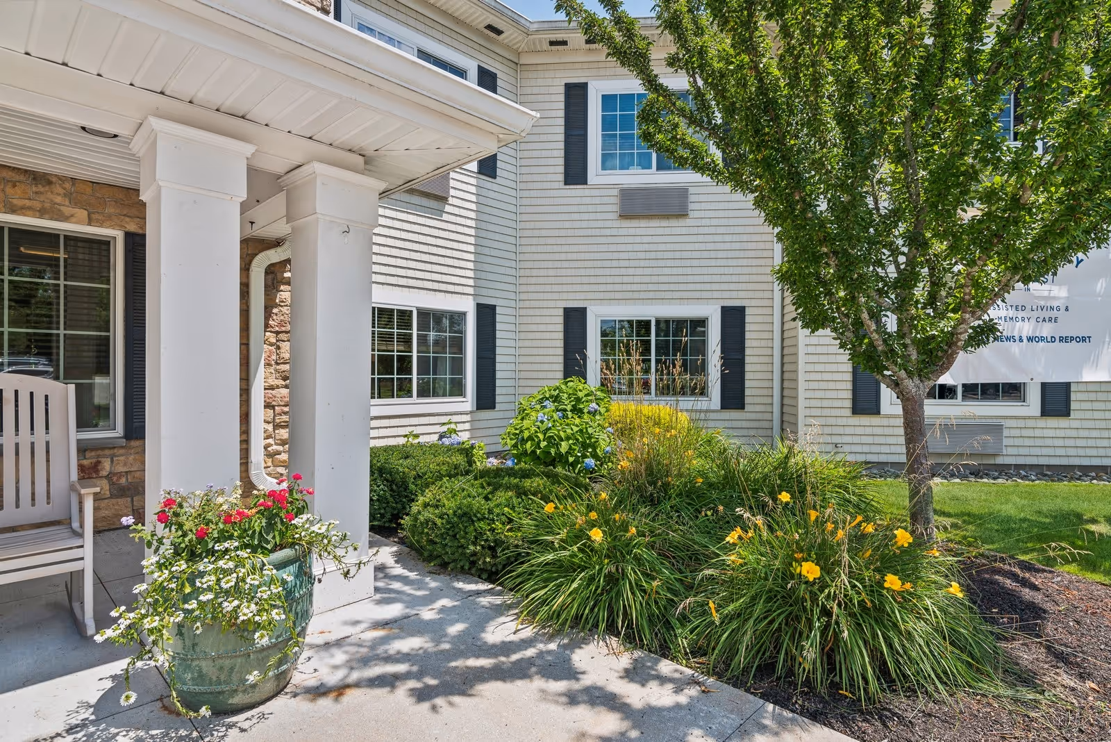 Outdoor view of the entrance area of a senior living facility with white pillars, a rocking chair, potted flowers, green bushes, yellow flowers, and a tree. The building has light-colored siding with black shutters on the windows.