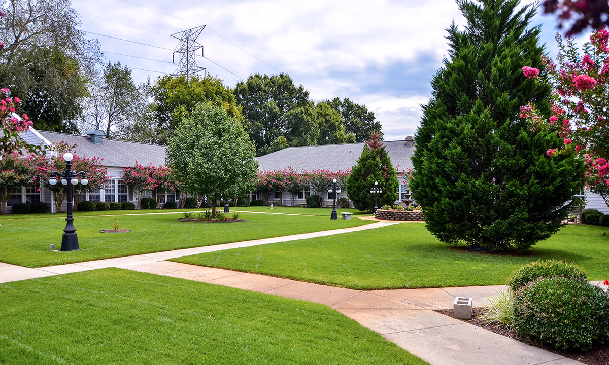 A well-maintained outdoor courtyard area with green grass, trees, flowering bushes, and paved walkways. The courtyard is surrounded by a single-story building with white siding and multiple windows. There are decorative black lamp posts and a partly cloudy sky overhead.