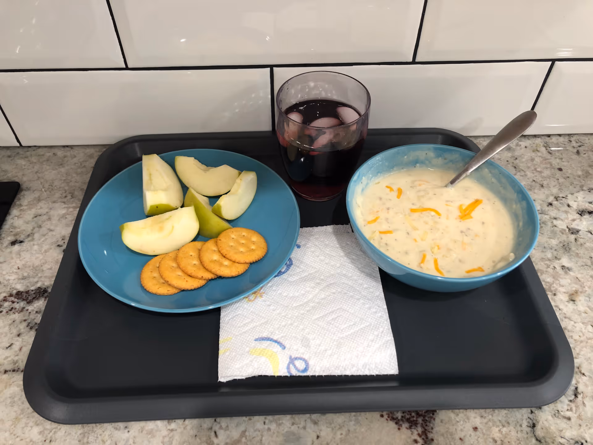 Tray on a kitchen counter with a bowl of creamy soup topped with cheese, a plate of apple slices and crackers, and a glass of red drink.