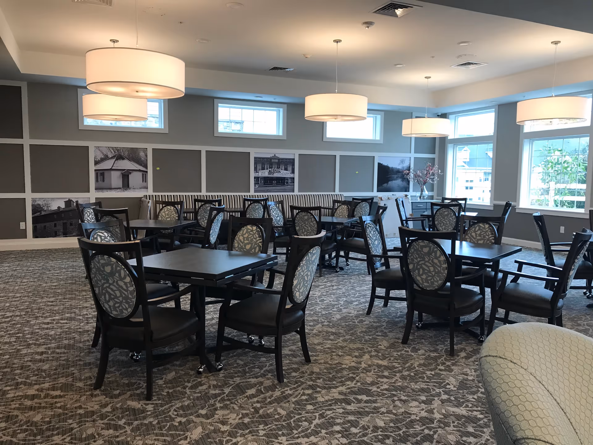 A spacious dining room with multiple square tables and cushioned chairs arranged neatly. The room features large windows allowing natural light to enter, modern pendant lights hanging from the ceiling, and a patterned carpet. The walls have decorative panels with black and white photos and a long striped bench along one side.