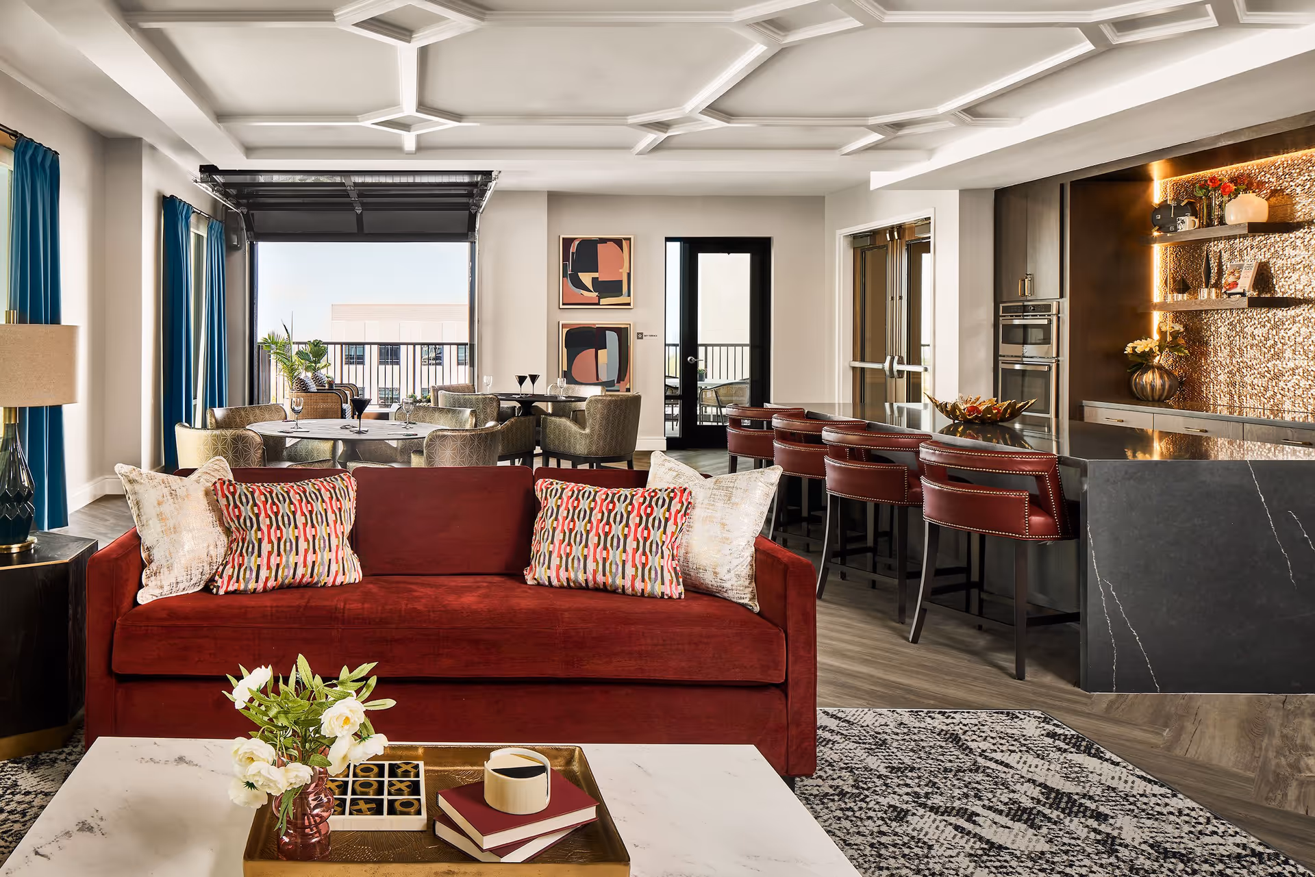 A stylish living room area with a red sofa adorned with patterned pillows, a white marble coffee table with a tray holding a vase of white flowers, books, and a cup. Behind the sofa is a dining area with round tables and upholstered chairs, and a kitchen bar with red leather stools and modern appliances. Large windows with blue curtains and a glass door lead to an outdoor balcony.