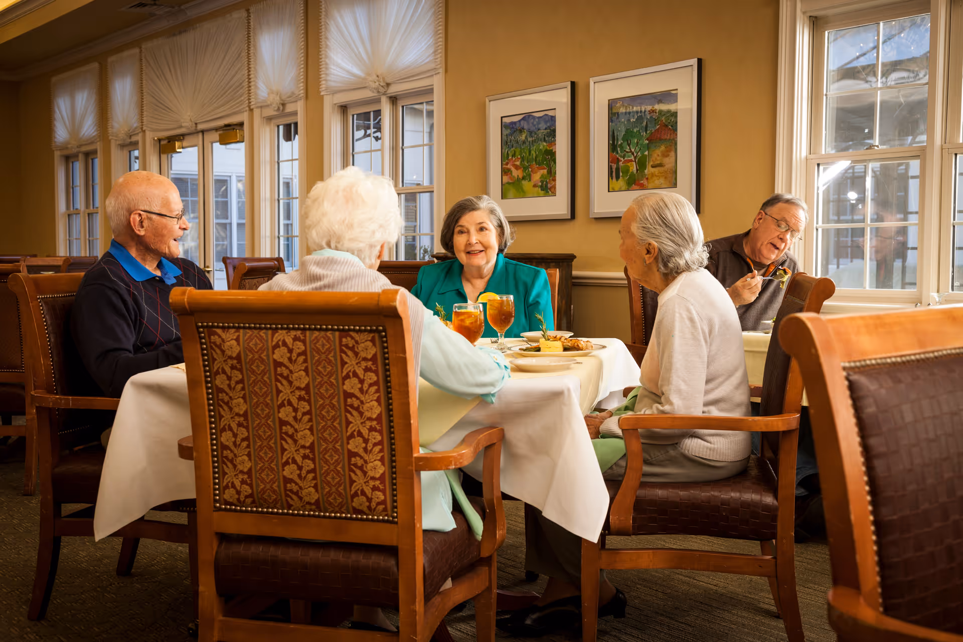 A group of elderly people sitting around a dining table in a well-lit room with large windows and framed artwork on the wall. They are enjoying a meal and drinks, engaging in conversation and smiling.