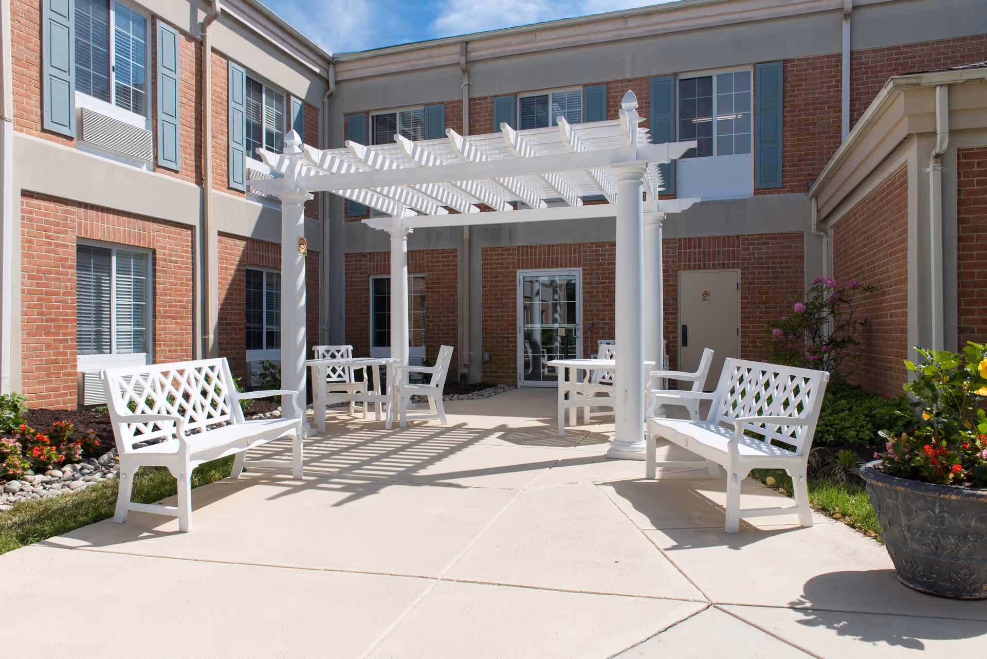 Outdoor courtyard area at Montcare Potomac featuring white benches and tables under a white pergola, surrounded by brick building walls with windows and some greenery including flowers and shrubs.