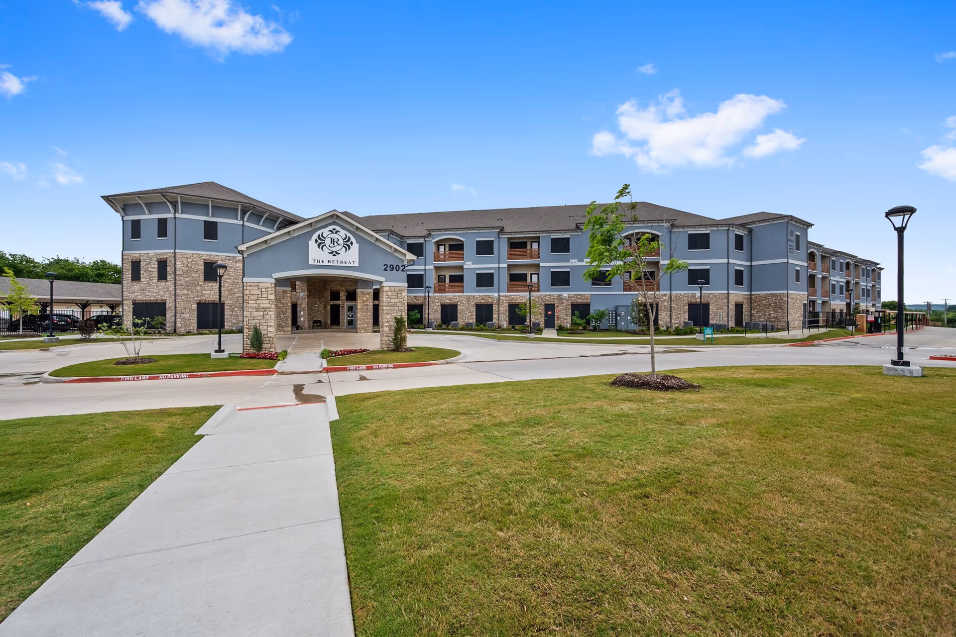 Exterior view of a large senior living facility building named The Retreat with a stone and blue-gray facade, multiple windows, and a covered entrance. The building is surrounded by a well-maintained lawn, a sidewalk leading to the entrance, street lamps, and a clear blue sky with a few clouds.