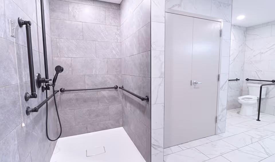A modern accessible bathroom with gray tiled walls and white tiled floor. The shower area has black grab bars and a handheld showerhead. There is a white toilet with black grab bars on the right side of the image, and a closed door in the center.