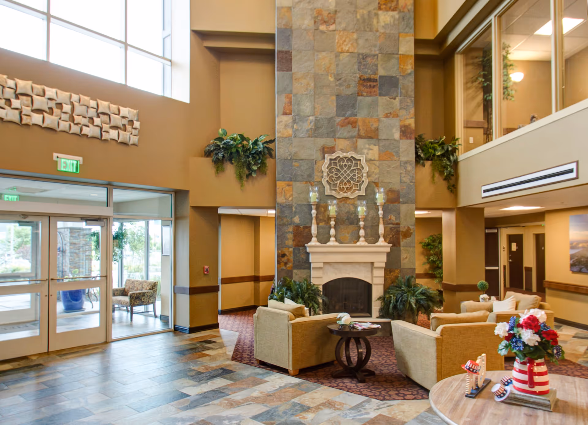 A spacious and well-lit living room area in a senior living facility with a tall stone fireplace as the focal point. The room features comfortable beige armchairs arranged around a small round table, decorative plants, and a large window letting in natural light. There is a round table with a red, white, and blue flower arrangement and small patriotic decorations in the foreground. The entrance doors and exit signs are visible on the left side.