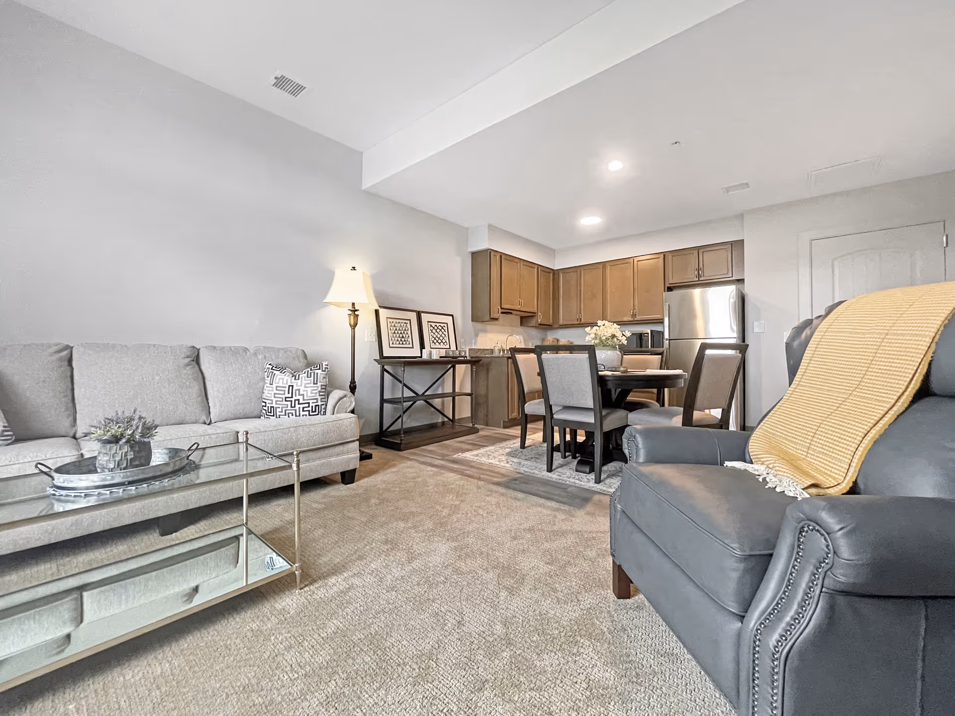 Open-concept living room and dining area with a gray sofa, leather armchair draped with a yellow throw, glass coffee table, round dining table, and a kitchen with wooden cabinets and a stainless refrigerator.