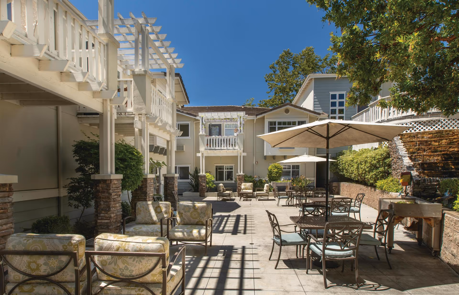 Outdoor patio area at Fremont Hills with cushioned chairs, tables with umbrellas, a stone water feature, and greenery under a clear blue sky.