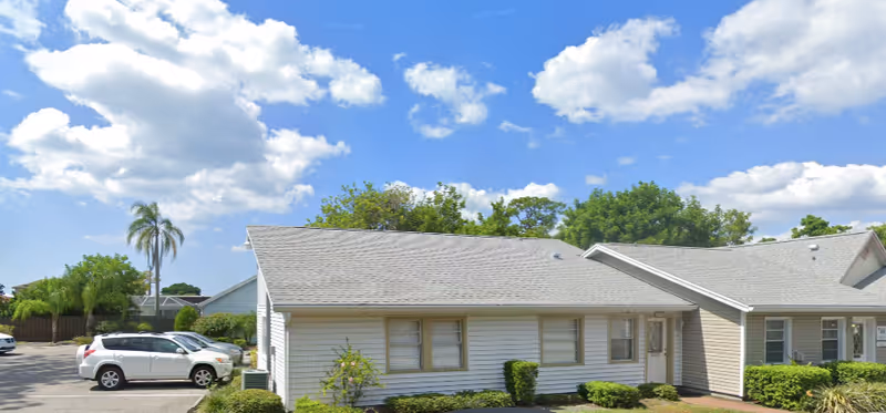 Exterior view of a single-story residential building with light-colored siding and a gray shingled roof under a blue sky with scattered clouds. There are several windows and a door visible on the front side of the building. In the foreground, there are green bushes and small plants along the building's foundation. To the left, a parking area with a few parked cars is visible, and a tall palm tree stands near the parking lot.