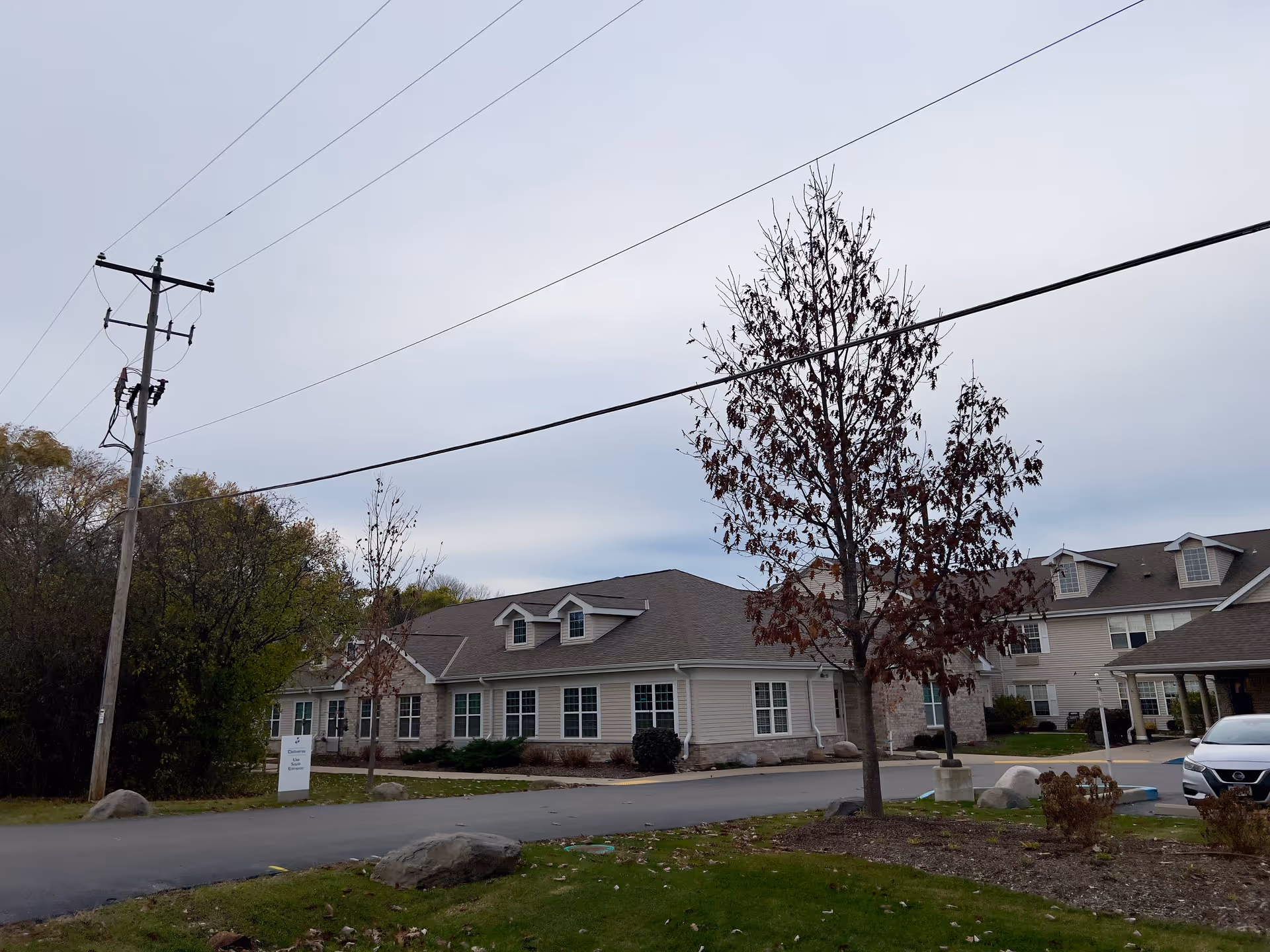 Exterior view of a senior living facility building with beige siding and stone accents, surrounded by trees and a driveway with a parked car. The sky is overcast.