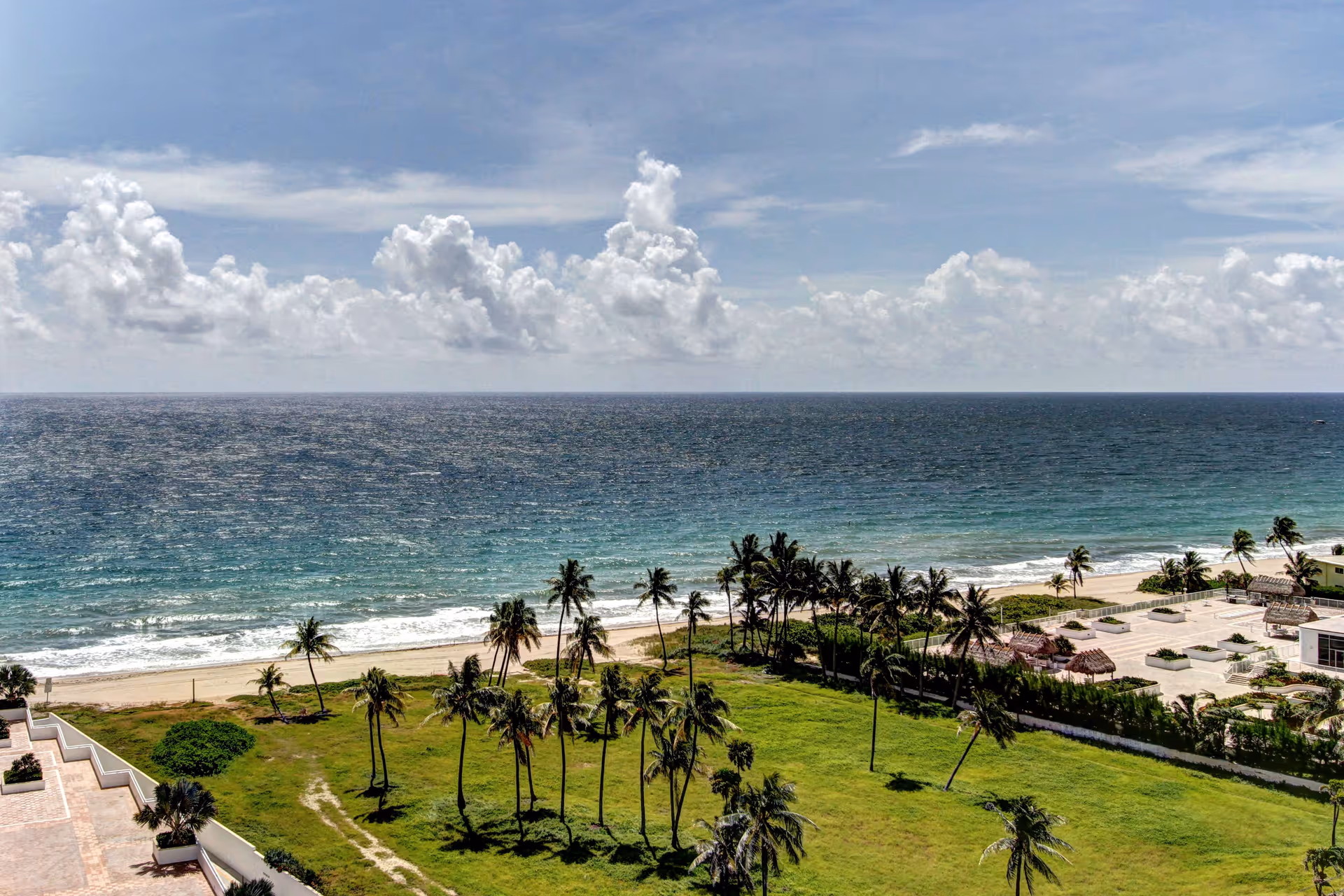 View of a beach with palm trees and green grass in the foreground, sandy shore, and the ocean under a partly cloudy sky.