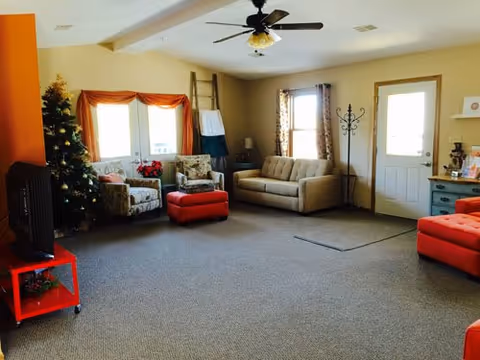 A cozy living room with beige walls and carpeted floor. The room features a ceiling fan, a beige sofa, two patterned armchairs, and two red ottomans. There is a Christmas tree decorated in the corner near a window with orange curtains. A TV sits on a red stand to the left, and a coat rack is near the white door on the right. A small blue cabinet with decorative items is also visible.
