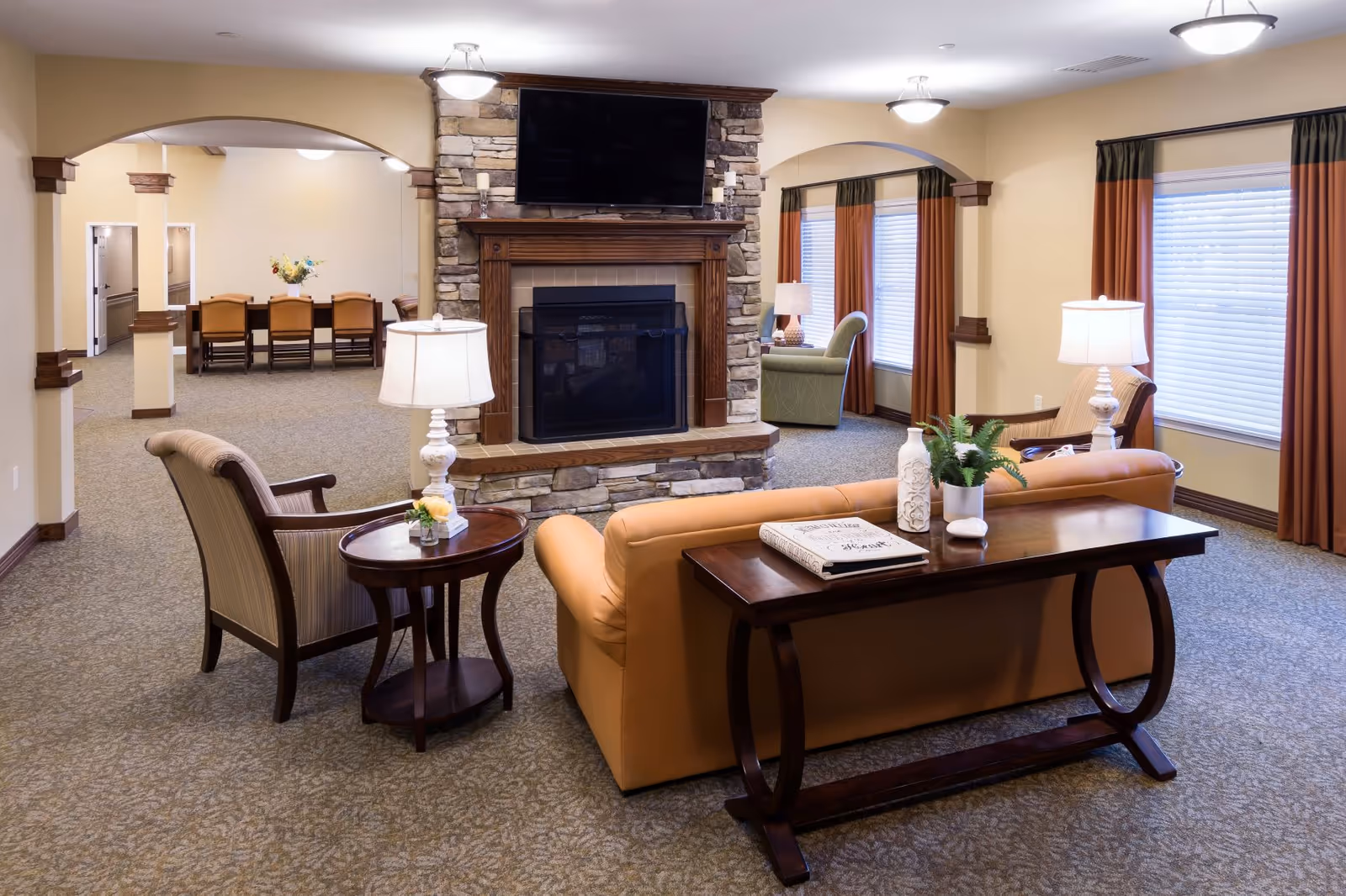 A cozy living room area in a senior living facility featuring a stone fireplace with a mounted TV above it. The room has a tan sofa with a wooden console table behind it, two armchairs, side tables with lamps, and large windows with brown and orange curtains. In the background, there is a dining area with a table and chairs and a flower arrangement on the table.