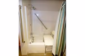 A bathroom with a white bathtub equipped with a showerhead and a grab bar on the wall. There is a white shower chair inside the tub and a shower curtain partially drawn to the side.