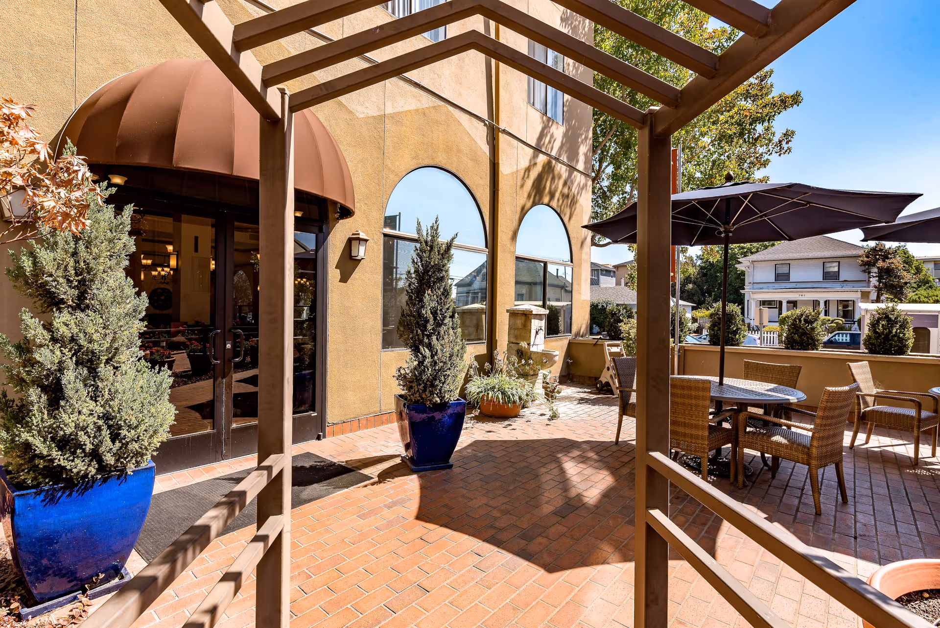 Outdoor patio area at San Leandro Senior Living with brick flooring, a pergola structure, potted plants, round tables with wicker chairs, and large umbrellas providing shade. The building exterior features arched windows and a brown awning over double glass doors.