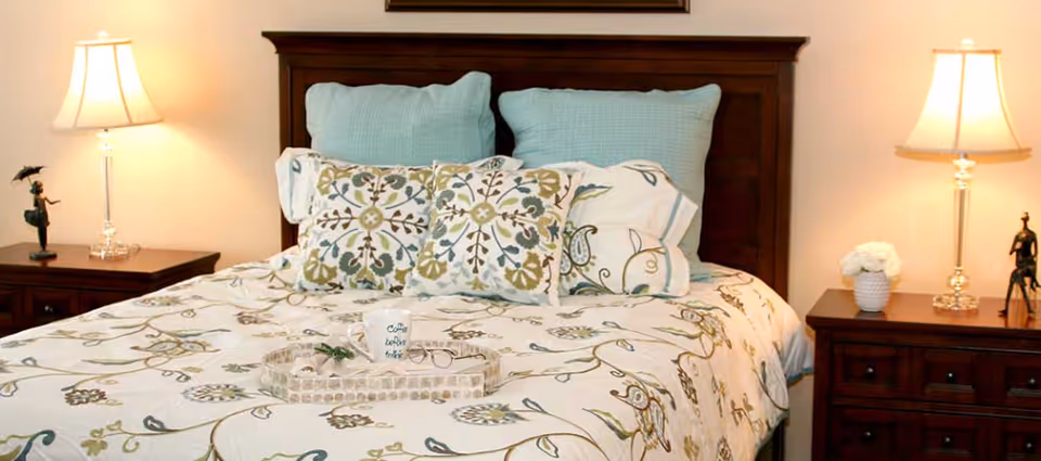 A neatly made bed with floral-patterned bedding, decorative pillows, a wooden headboard and two nightstands topped with lamps.