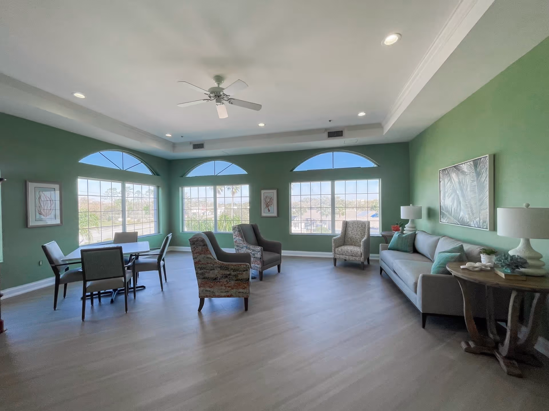 A spacious, well-lit common area with green walls and large windows featuring arched transoms. The room contains a gray sofa with pillows, several armchairs, a round wooden side table with a lamp and decorative items, and a small dining table with four chairs. The ceiling has recessed lighting and a white ceiling fan.