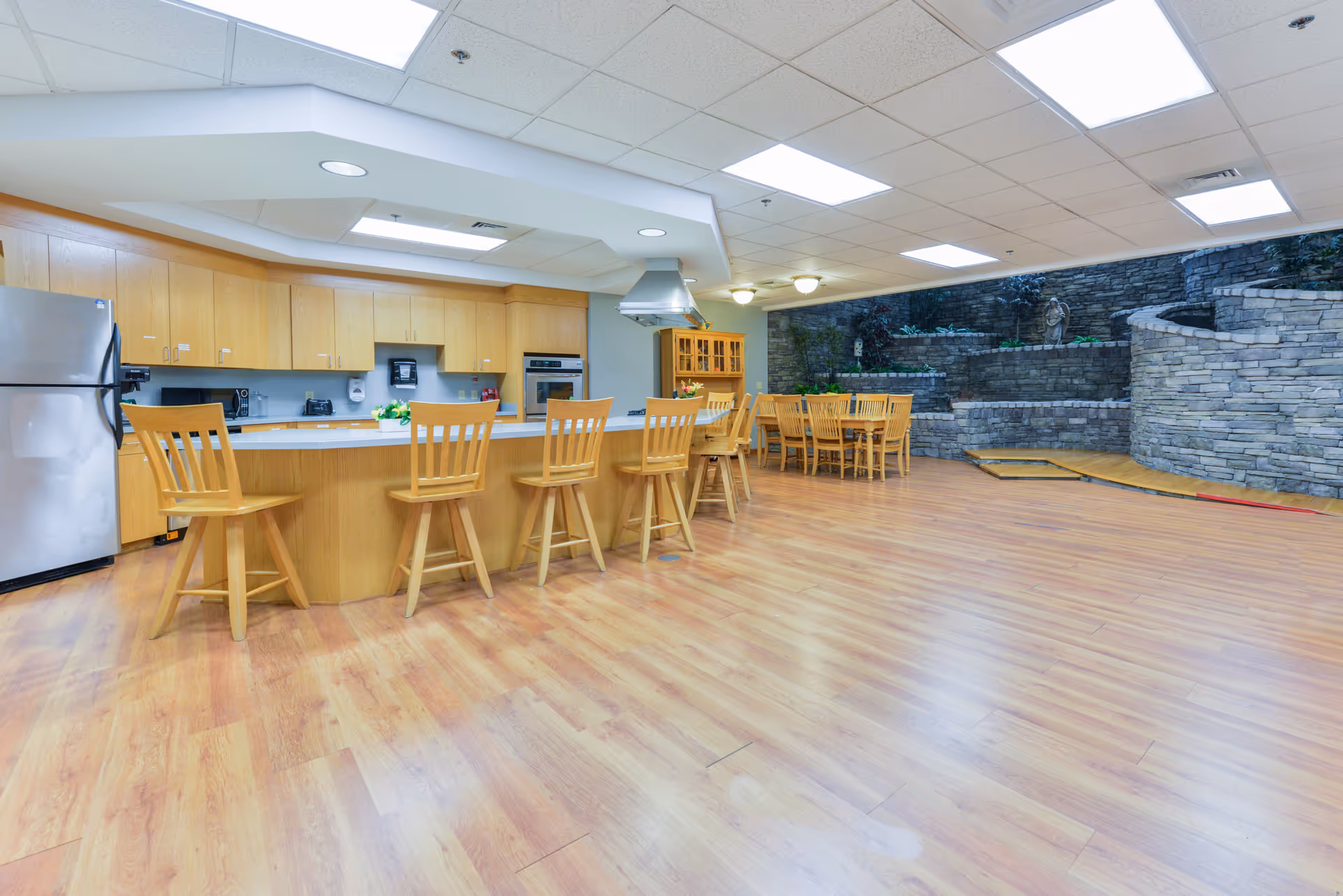 A spacious kitchen and dining area with wooden flooring and light wood cabinetry. The kitchen features a large island with five wooden bar stools, a stainless steel refrigerator, built-in oven, and a range hood. In the background, there is a dining table with chairs and a stone wall with plants and a small statue, creating a cozy atmosphere.