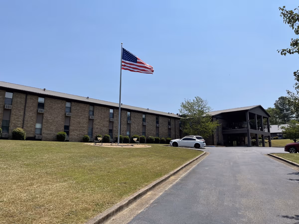 Exterior view of a two-story senior living facility building with a large American flag on a flagpole in front. There is a driveway leading up to the building entrance, with a white car parked near the entrance and some trees and bushes around the building under a clear blue sky.