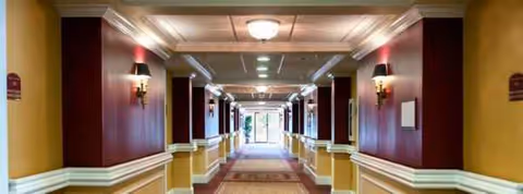Long, well-lit hallway with maroon walls, lamps, and carpet in a senior living facility.