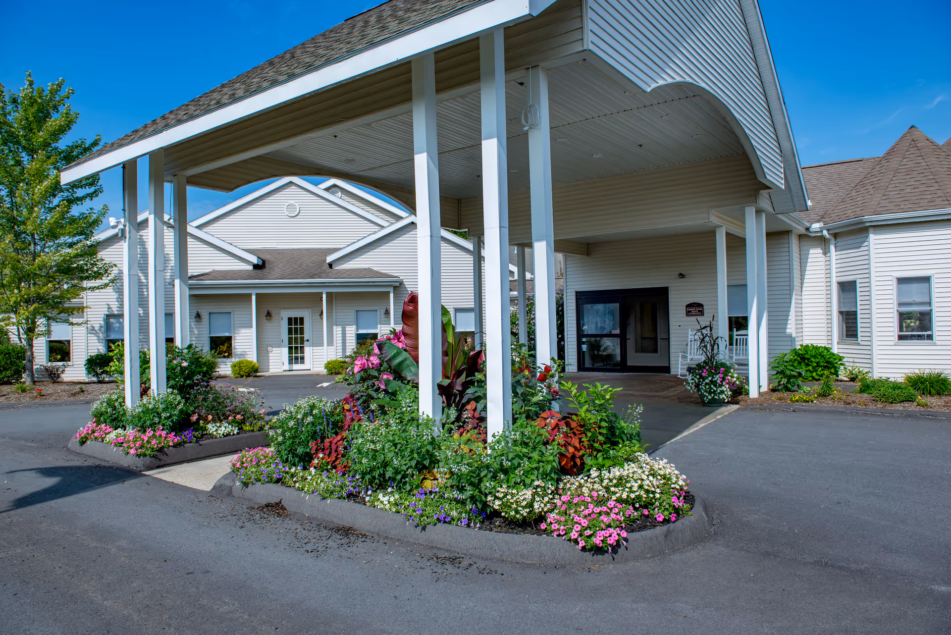 Entrance of a senior living facility with a covered driveway supported by white pillars. The area around the driveway is landscaped with colorful flowers and green plants. The building is light-colored with multiple windows and a clear blue sky overhead.