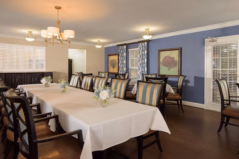 Well-lit dining room with long tables covered in white tablecloths, floral centerpieces, and striped upholstered chairs.