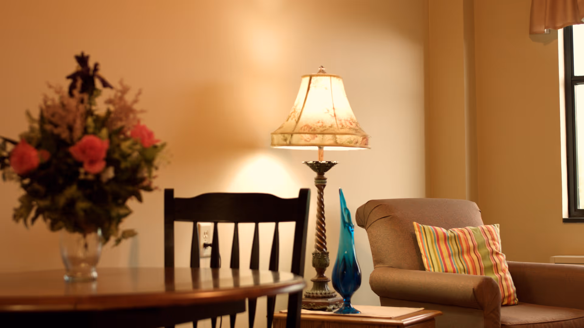 A cozy interior scene featuring a wooden table with a vase of flowers, a dark wooden chair, a decorative table lamp with a floral lampshade, a blue glass ornament, and a comfortable armchair with a striped cushion near a window.