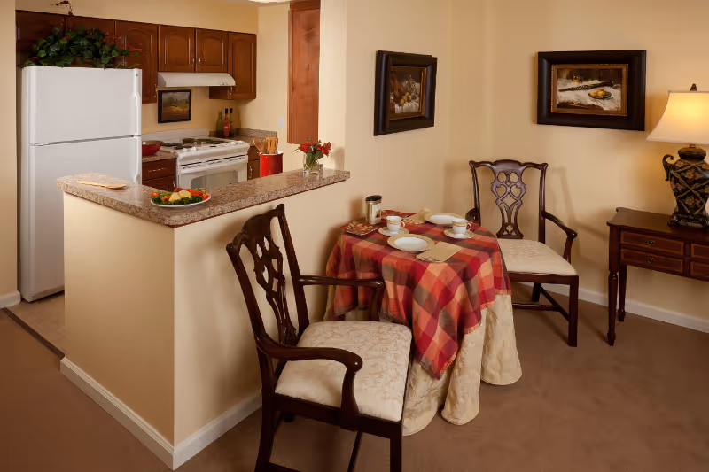 Small dining area with a round table set for two and two chairs next to an open kitchenette with a refrigerator and stove.