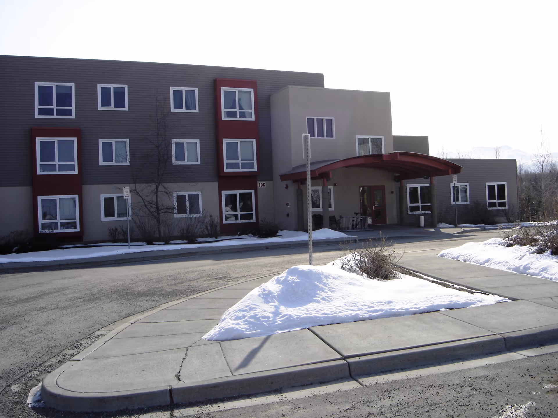 Three-story senior living building front with a red covered entrance, snow on the sidewalks and a circular driveway.