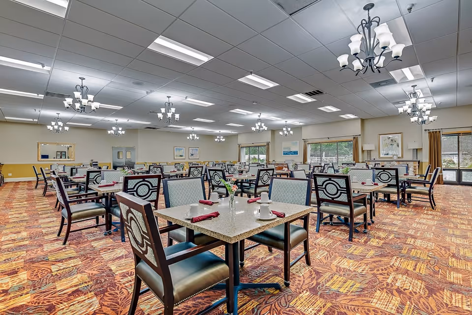 Spacious dining room with rows of set tables and chairs, chandeliers, large windows, and patterned carpet.